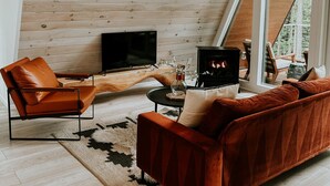 A modern loft-style living room with a sloped ceiling, wooden paneling, a large TV, a fireplace, and a cozy seating area. - Fox Ridge-Black Alder Lodging (Logan)