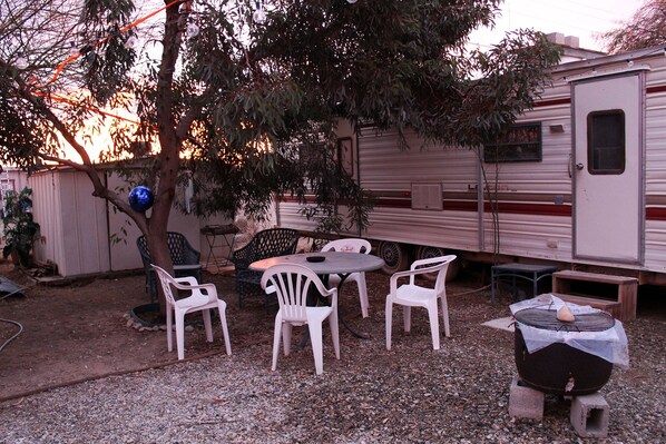 A recreational area with a table and chairs, a barbecue grill, and a mobile home in the background. - Cozy 80s Safari Style RV in Tranquil Bombay Beach with AC (BOMBAY BEACH)