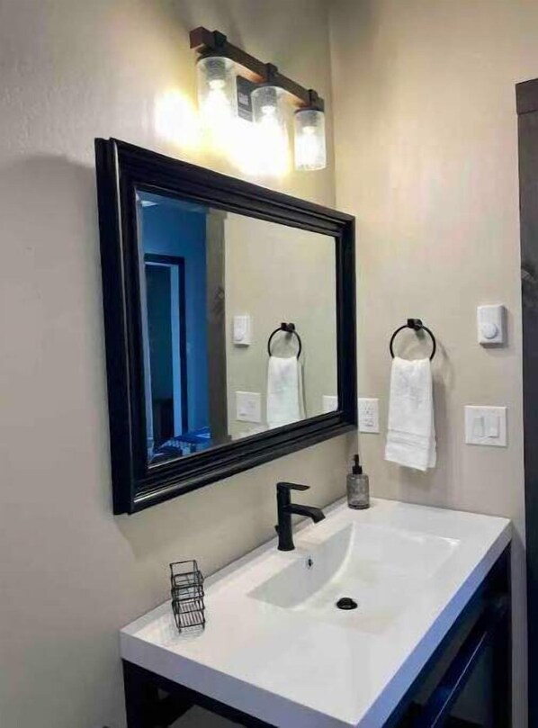 Une salle de bain avec un lavabo blanc, un robinet noir et un grand miroir surmonté de trois lampes.