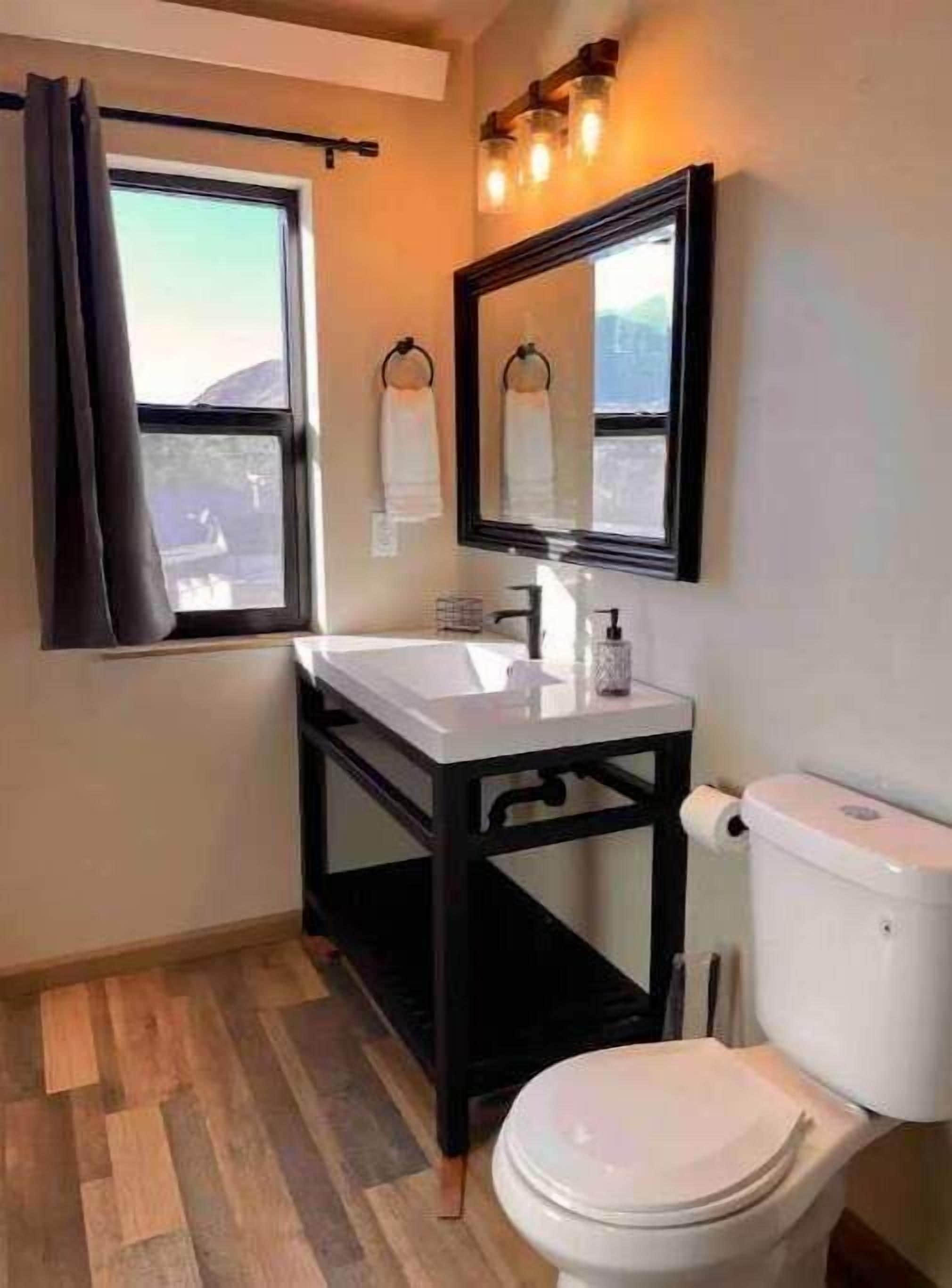 A bathroom with a white toilet, a sink with a black frame, a window with a curtain, and a mirror above the sink.