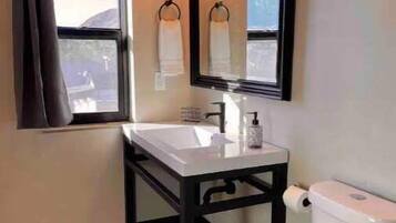 A bathroom with a white toilet, a sink with a black frame, a window with a curtain, and a mirror above the sink.