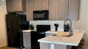 Cuina compacta amb armaris de fusta, un refrigerador negre, un microones i una cuina.