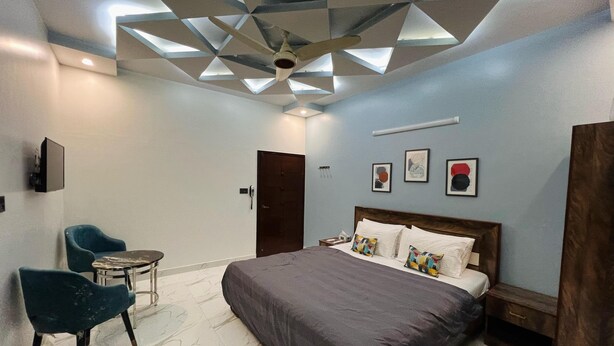 Un dormitorio moderno con cama, escritorio, dos sillas, ventilador de techo y una obra de arte en la pared.