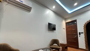 Una habitación con piso de madera, una mesa con cubierta de vidrio, dos sillones tapizados, un televisor montado en la pared y una puerta de madera con arco de ladrillo.