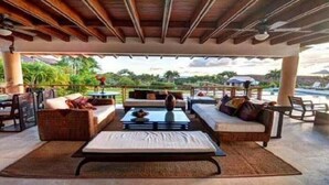 Un espacio de estar al aire libre cubierto con sofás, una mesa de centro y vistas a una piscina y follaje.