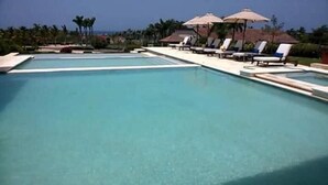 Una piscina con sillas de descanso y sombrillas, rodeada de palmeras y un cielo despejado.