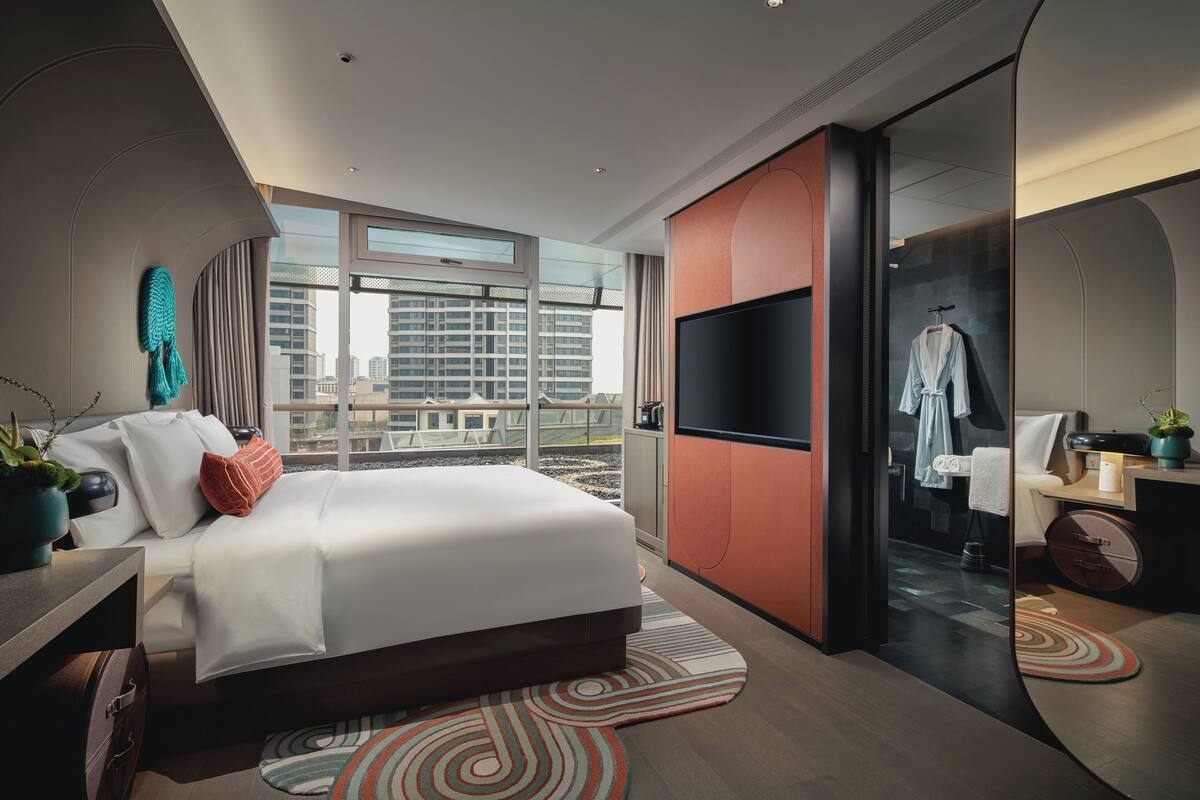 A modern hotel room with a large bed, a TV, a mirror, and a view of the city through large windows.