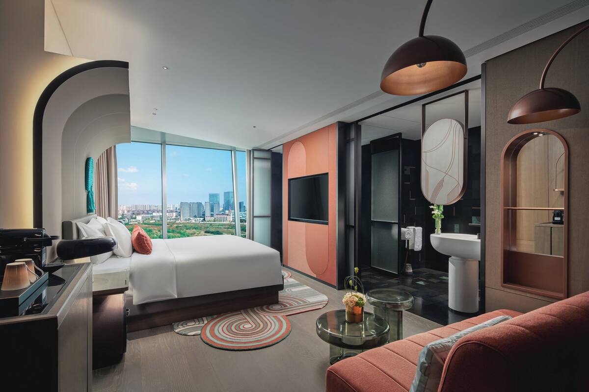 A modern hotel room with a large bed, a desk, a sofa, a glass coffee table, and a view of the city.