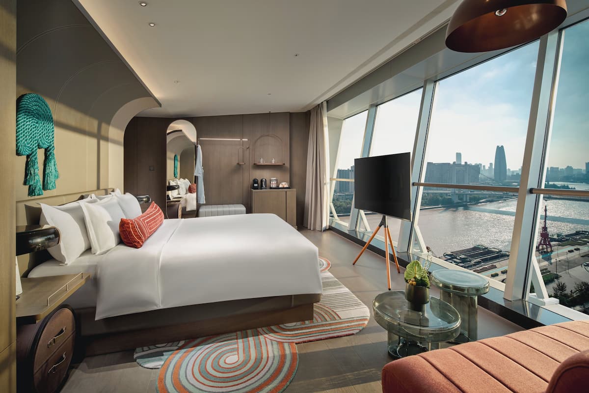 A modern hotel room with a large bed, a TV, a balcony with a city view, and a small seating area.
