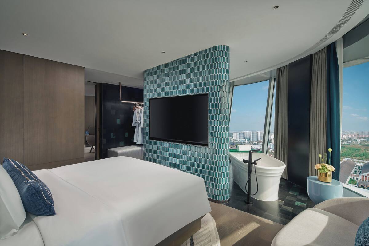 A modern hotel room with a large bed, a flat-screen TV mounted on a tiled accent wall, a small round table with a vase of flowers, and a view of the city through large windows.