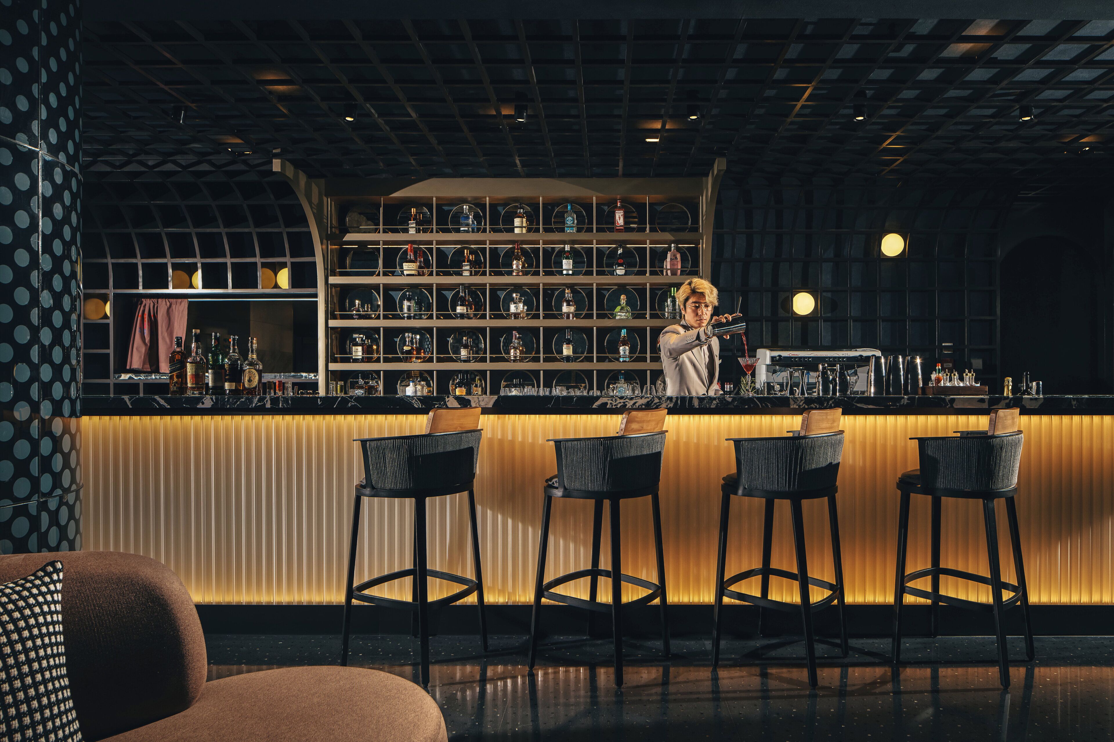 Una zona de bar con mostrador, taburetes y estanterías llenas de botellas.