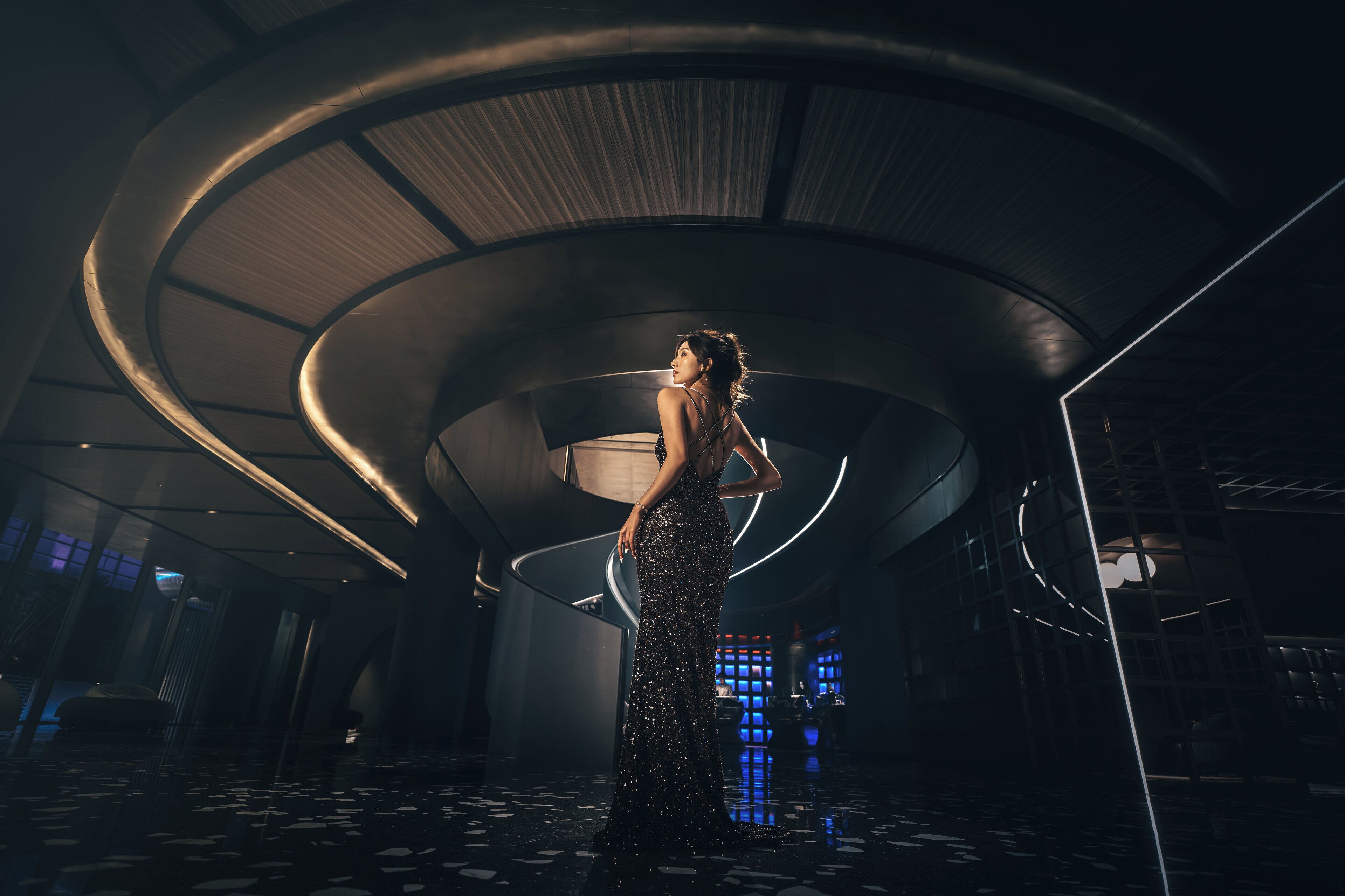 Una mujer con un vestido largo, oscuro y con lentejuelas posa en un vestíbulo moderno de diseño circular, con techo alto y suelo reflectante.