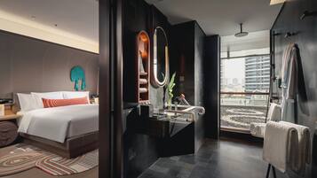 Una habitación de hotel moderna con una cama grande, un tocador con espejo, zona de ducha y vistas a la ciudad desde la ventana.