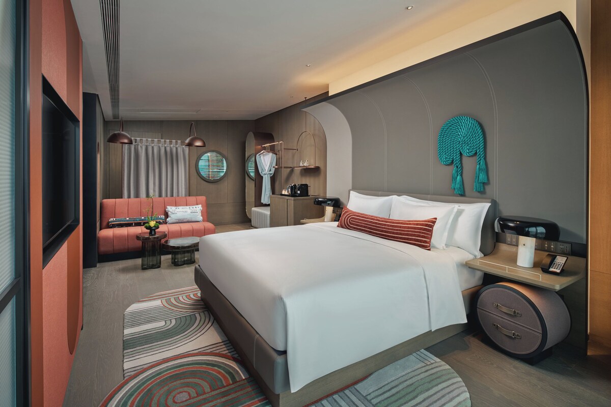 Habitació d'hotel moderna amb un llit gran, un sofà, una taula de treball amb una làmpada i una televisió.