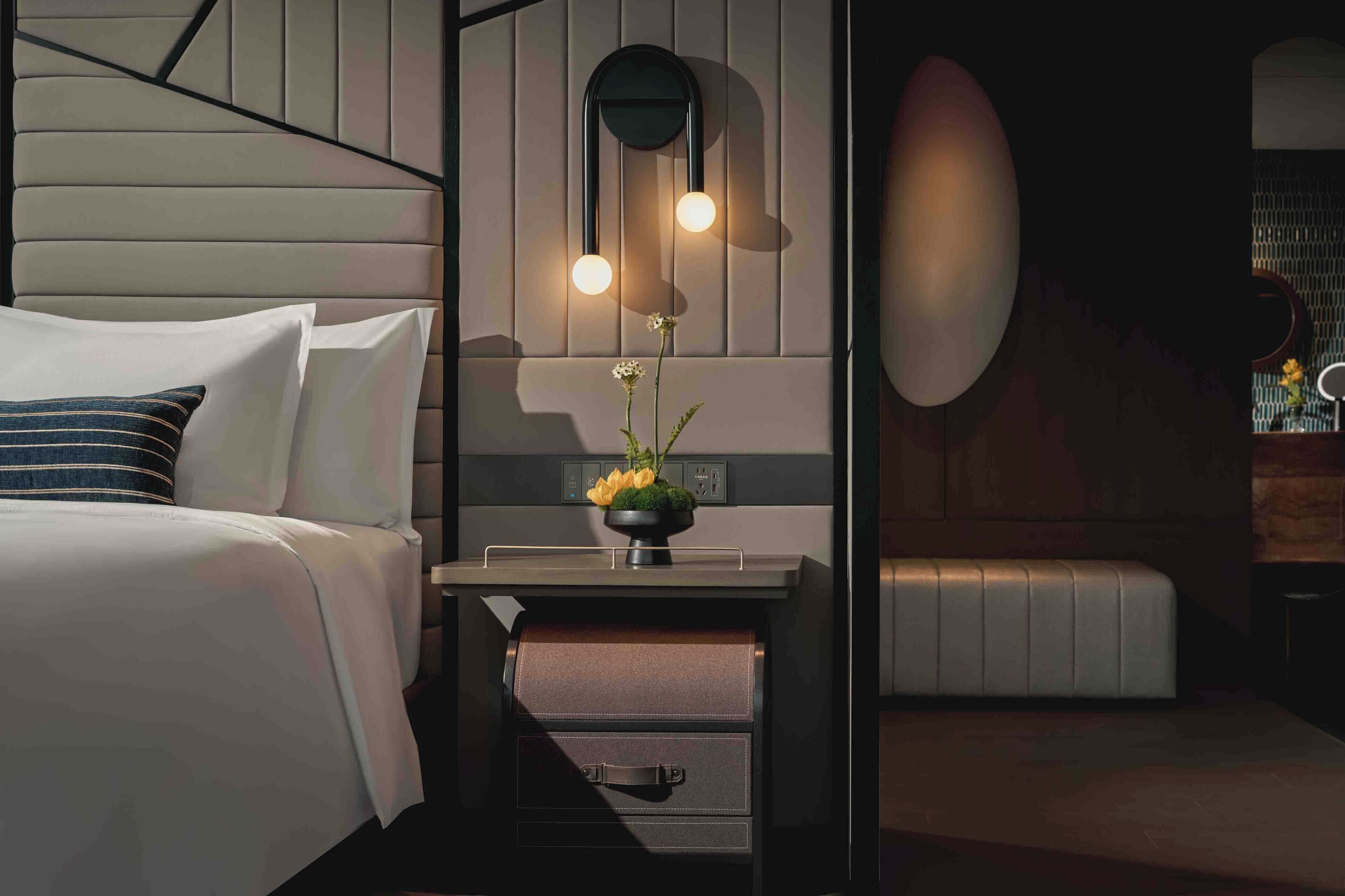A modern hotel room with a bed, bedside table, and a small desk area with a lamp and a vase of flowers.