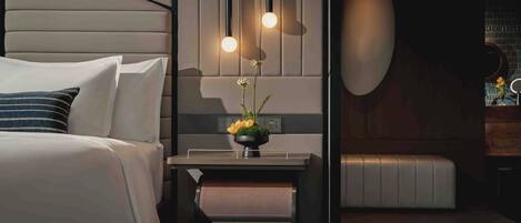 A modern hotel room with a bed, bedside table, and a small desk area with a lamp and a vase of flowers.