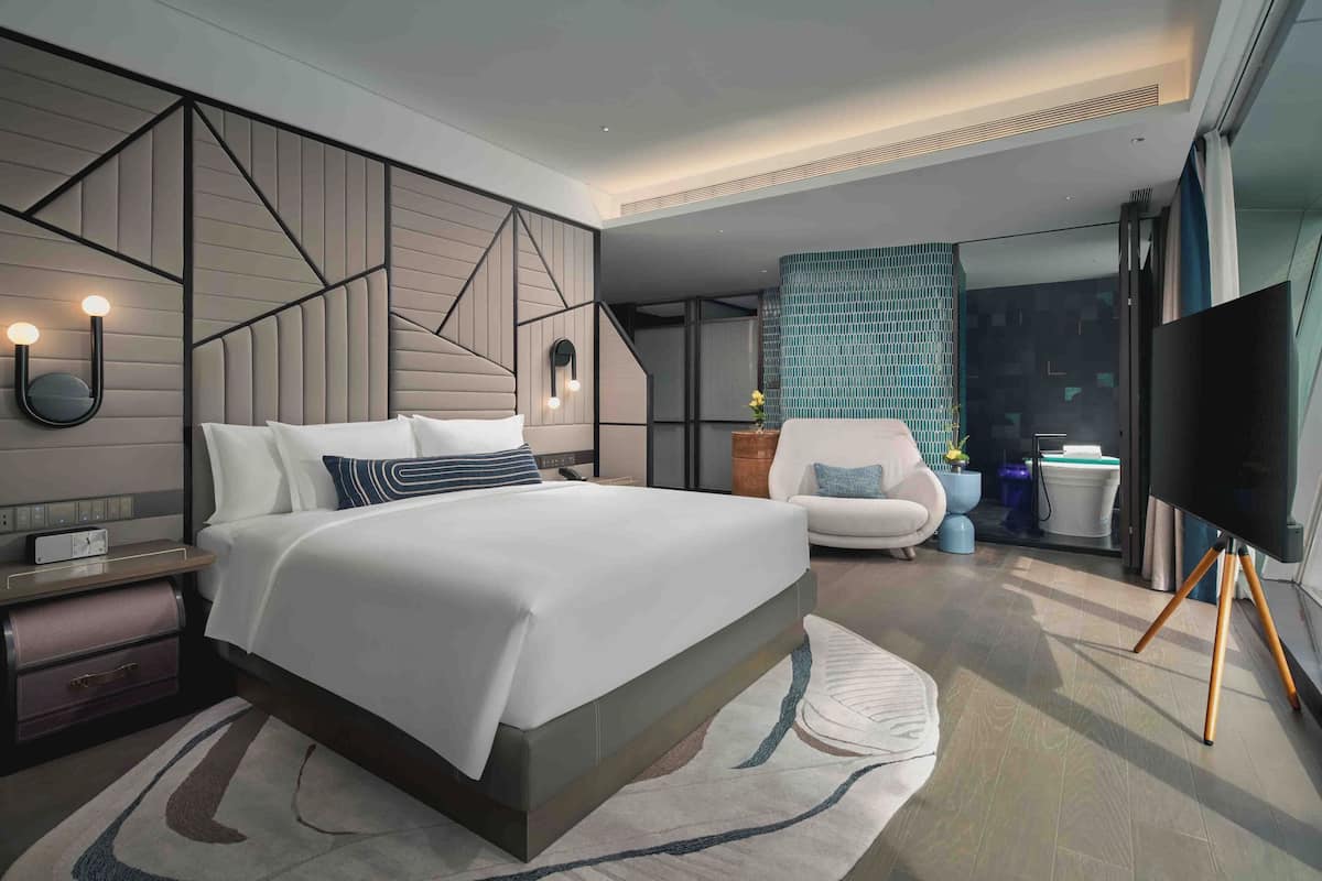 A modern hotel room with a large bed, a flat-screen TV, a seating area with a chair and side table, and a decorative wall panel.
