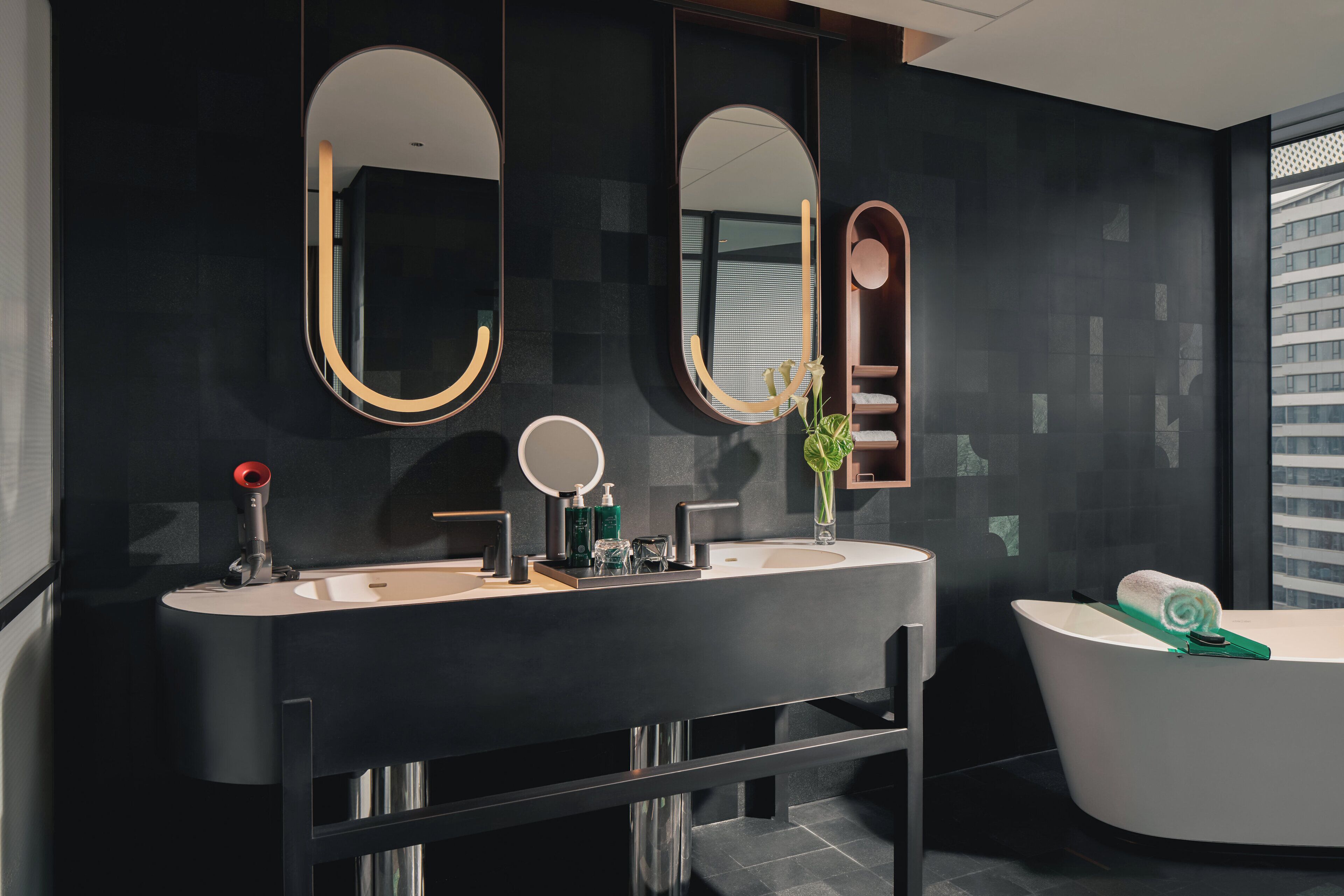 A modern bathroom with a large vanity, two mirrors, a bathtub, and a window with a city view.