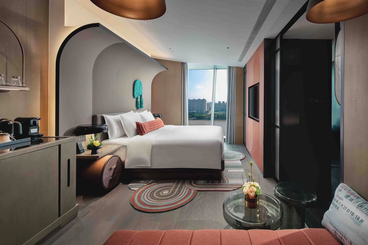 A modern hotel room with a bed, a desk with a coffee maker, a small table with a vase, and a large window offering a city view.