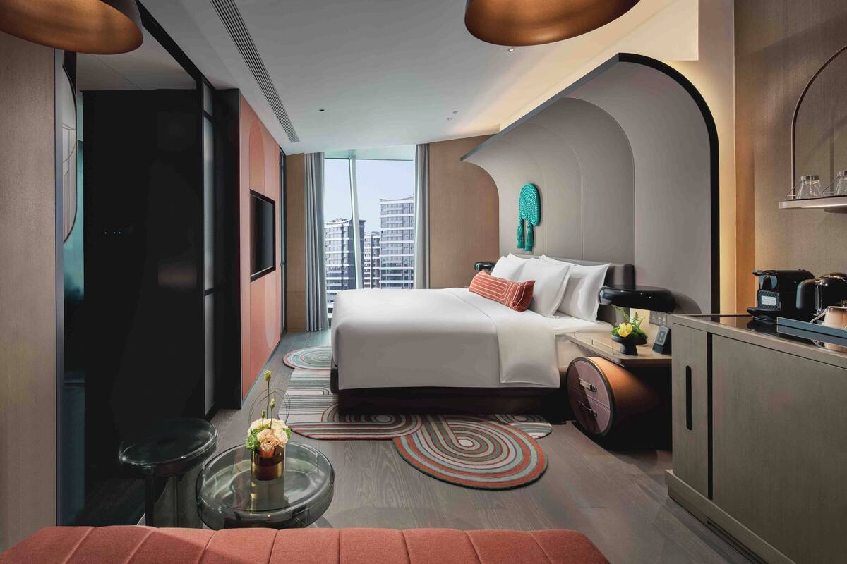 A modern hotel room with a large bed, a desk with a coffee machine, a glass table with a vase, and a view of the city.