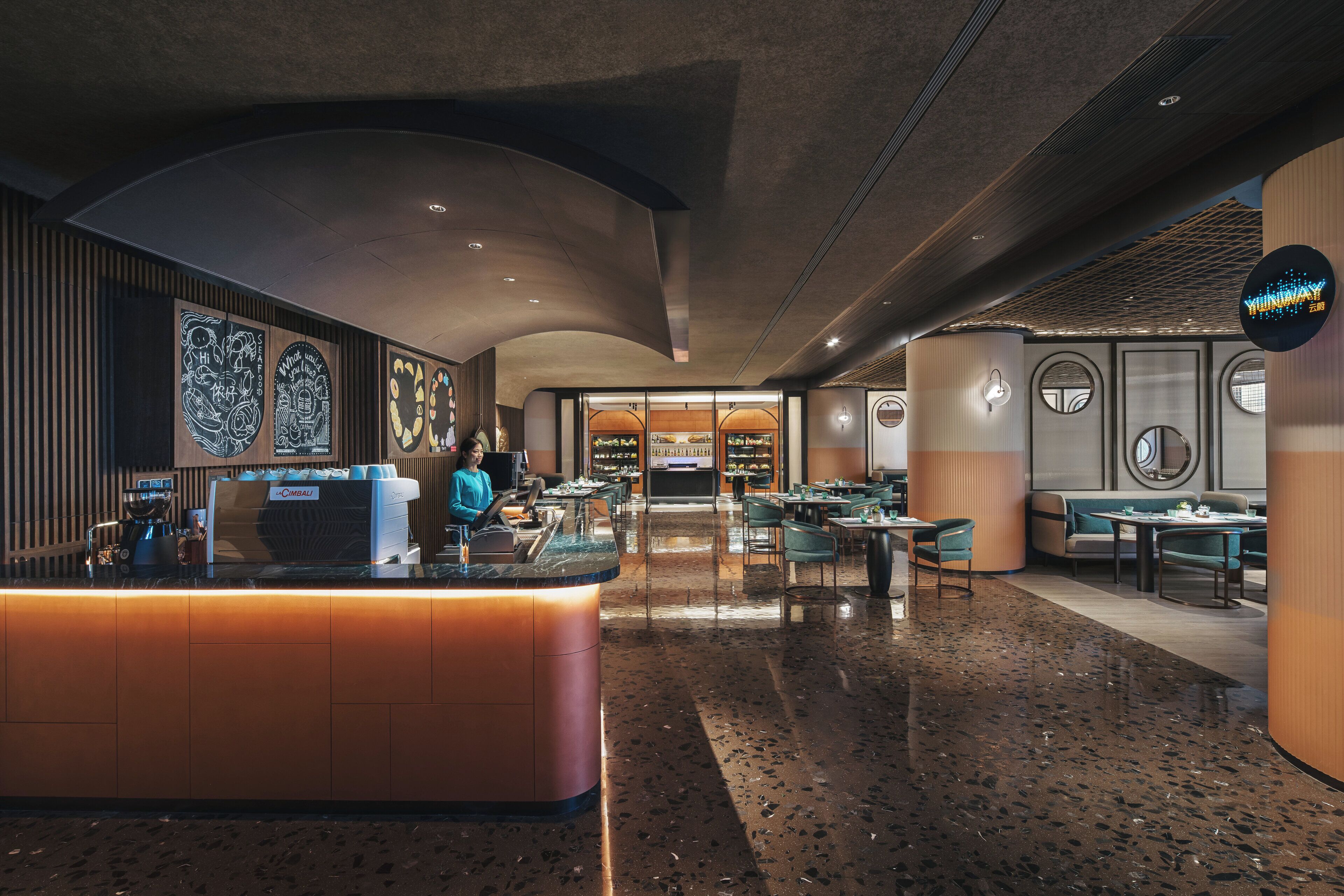 Una moderna hall d'albergo con una reception centrale, zone salotto e vista sul ristorante sullo sfondo.
