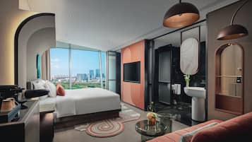 A modern hotel room with a large bed, a desk, a sofa, a glass coffee table, and a view of the city.