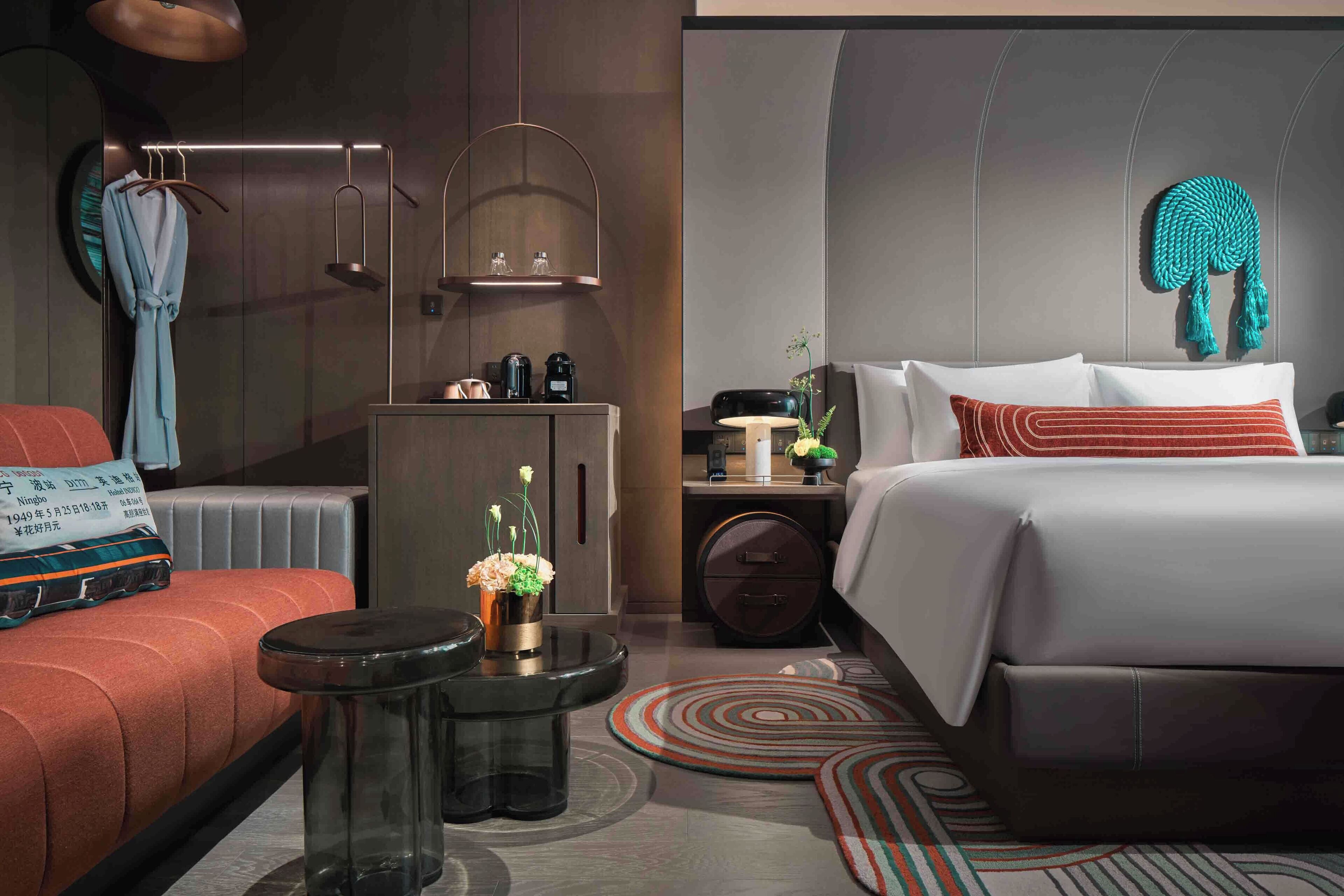 Habitación de hotel moderna con cama, zona de estar con sofá rojo, pequeña mesa con un jarrón de flores, mesita de noche con lámpara y un adorno en la pared.