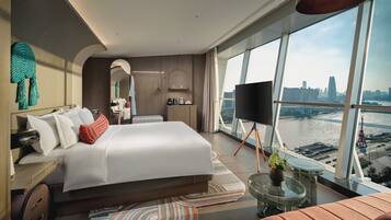 A modern hotel room with a large bed, a TV, a balcony with a city view, and a small seating area.