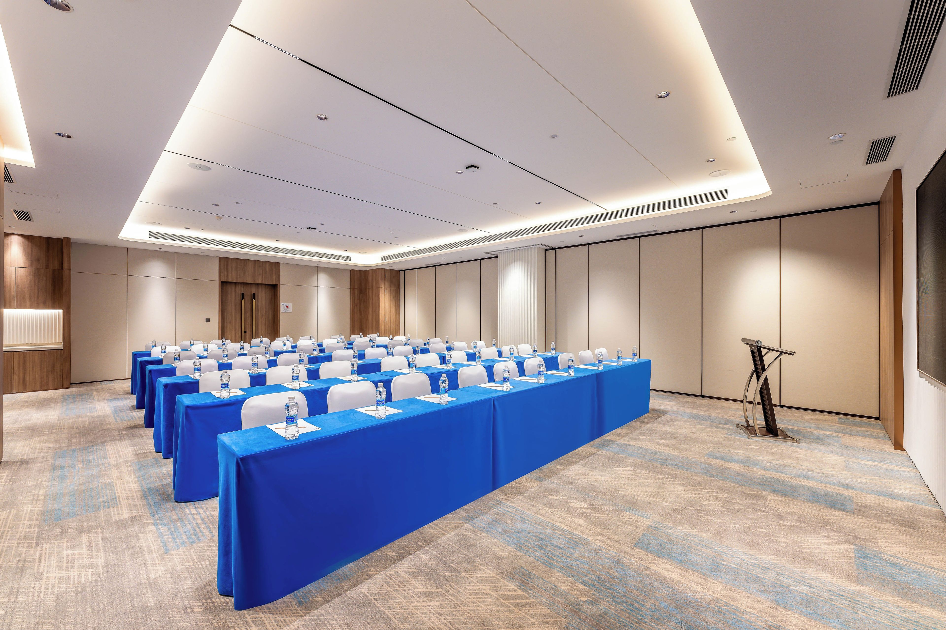 Un salón de conferencias con filas de mesas, cada una con mantel azul y botellas de agua.