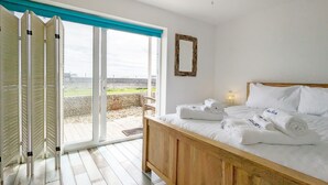 A bedroom with a wooden bed, white linens, a ceiling fan, and a view of a grassy area through a sliding glass door. - Sea Esta, Selsey - sleeps 4 guests  in 1 bedroom (Selsey)
