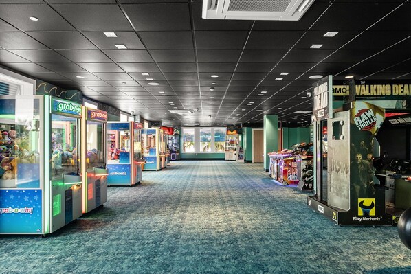 An indoor arcade with claw machines and gaming stations, featuring 'The Walking Dead' and 'Raw Thrills' game machines.