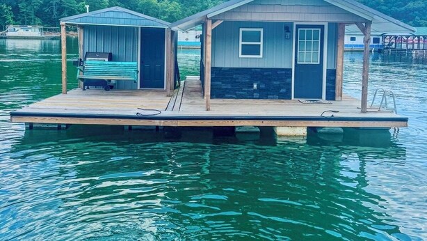 Une maison flottante avec un quai, un espace extérieur aménagé et couvert, et un petit bateau.