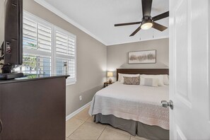 Une chambre à coucher avec un lit, un ventilateur de plafond, une table de chevet, une lampe et une fenêtre avec des stores.