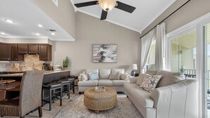 A modern kitchen with dark wooden cabinets, a dining area with a wicker chair, a round woven ottoman, and a beige sofa in a living room with a large window. - 10G, Sandy´s Beach Retreat (Destin)