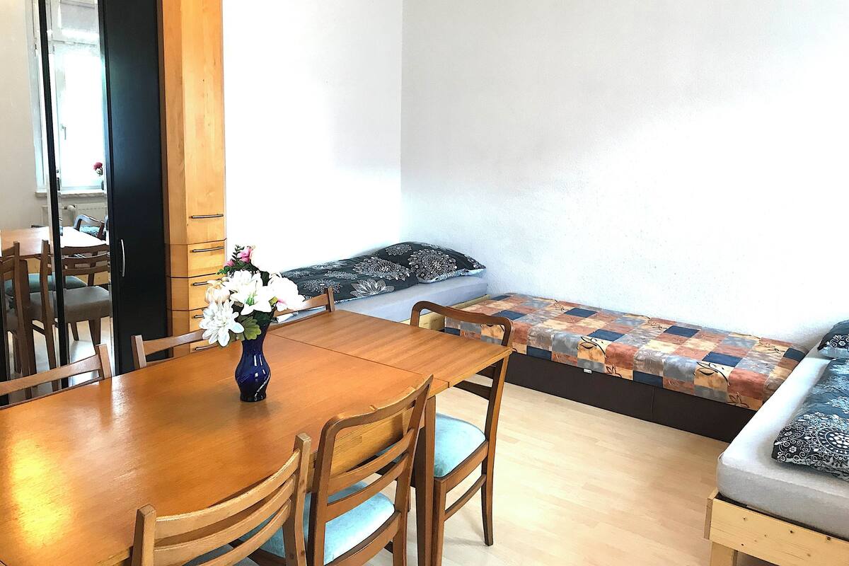 A dining area with a wooden table and chairs, a vase with flowers, and a sofa with patterned cushions.
