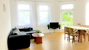 A modern living room with a black sofa, a small coffee table with a plant, a flat-screen TV, and a dining area with a table and chairs. - Meeraner Parkresidenz (Meerane)