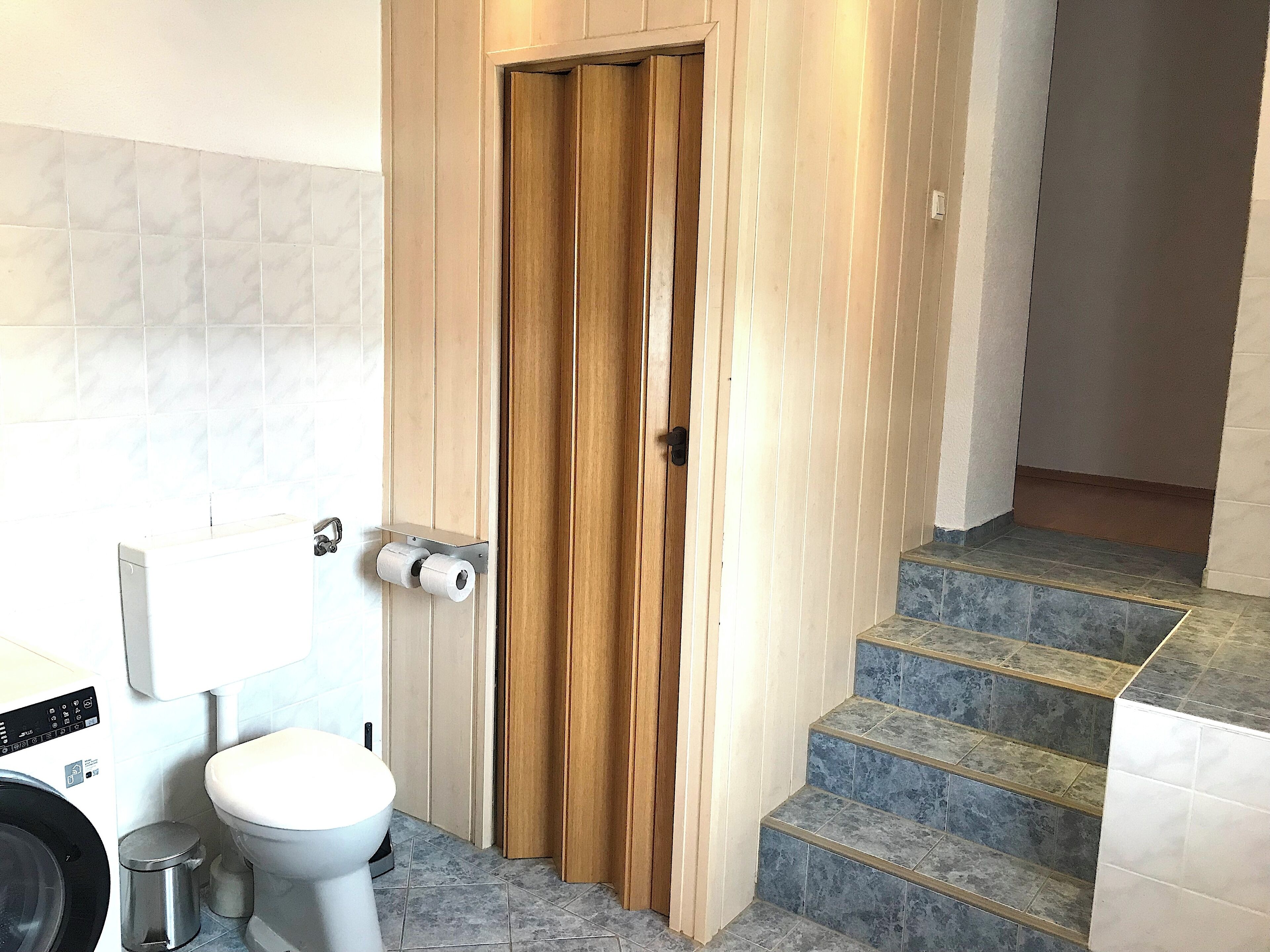A bathroom with a toilet, a paper towel dispenser, a washing machine, and a trash bin.