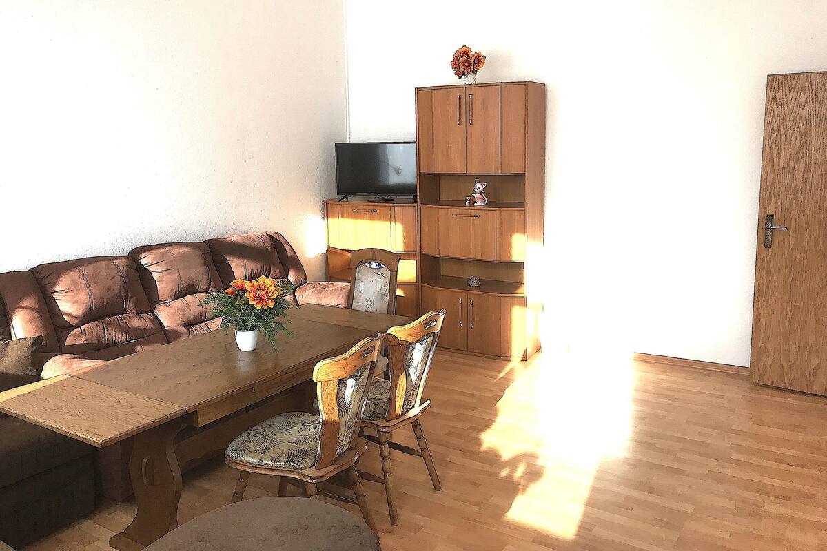A living room with a wooden sofa, a dining table with chairs, a TV on a cabinet, and a door.