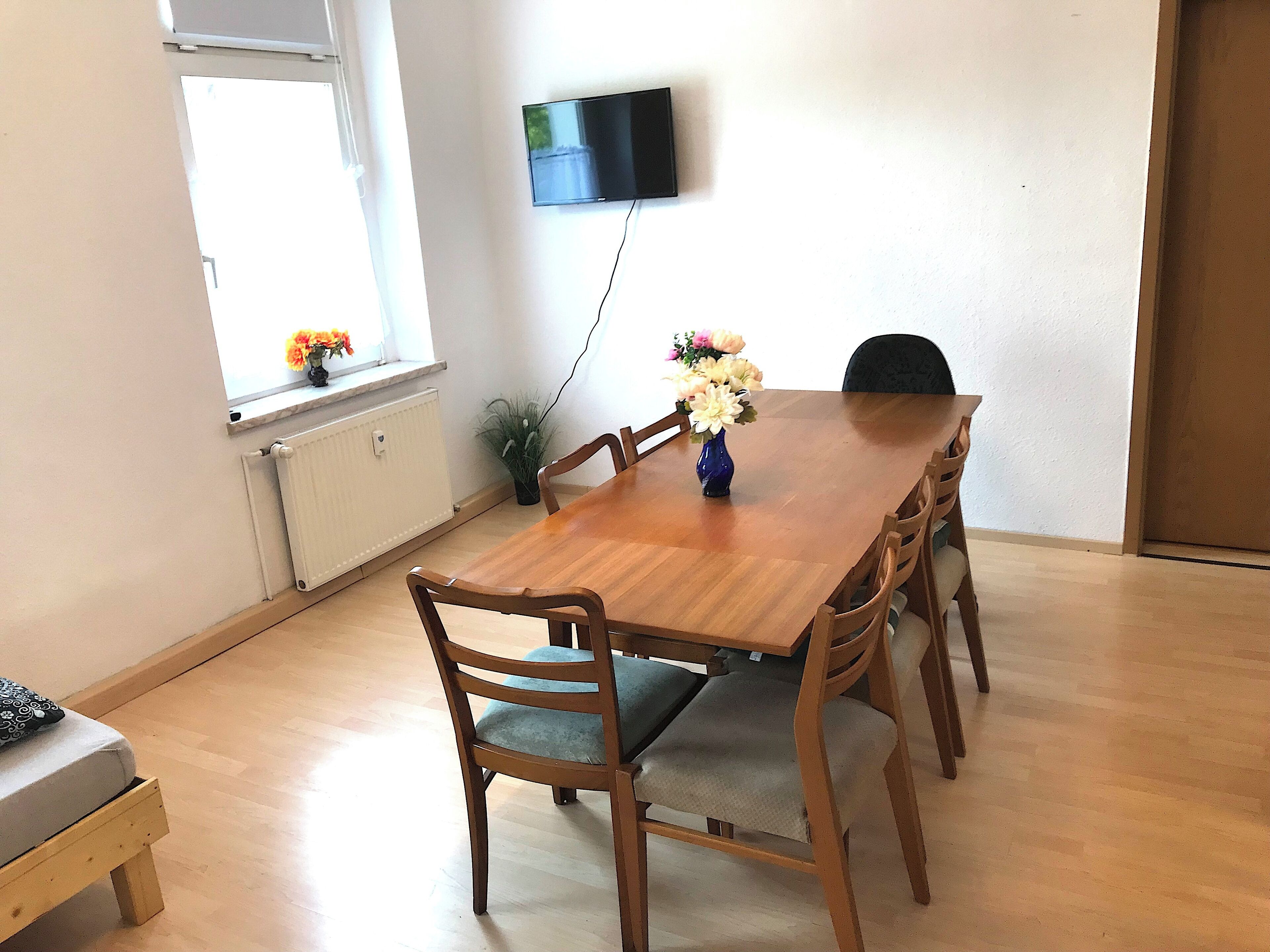 A dining area with a wooden table, six chairs, a vase with flowers, a television on the wall, and a window with flowers.