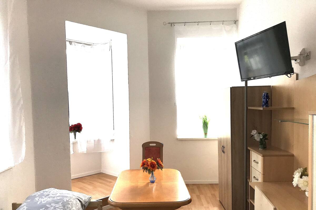 A small, well-lit room with a wooden dining table, a single chair, a TV mounted on the wall, and a window with a curtain.