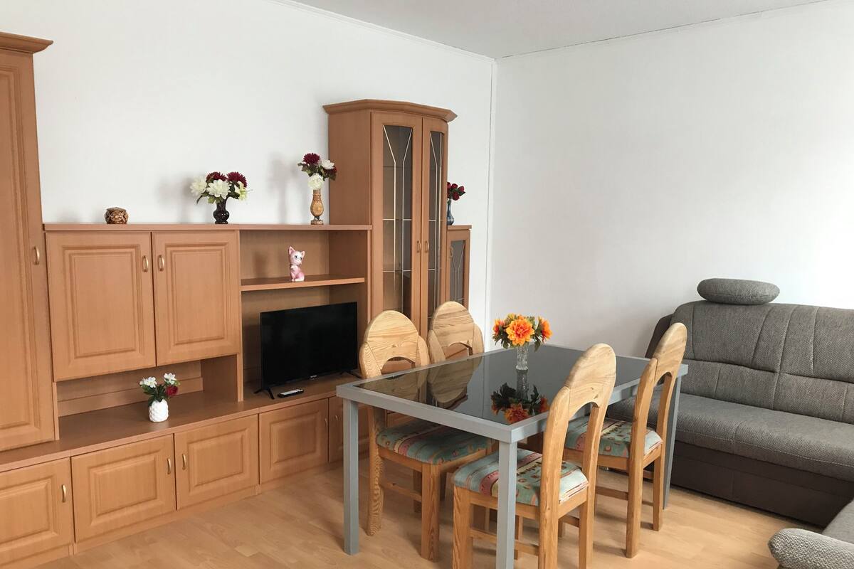 A living room with a glass dining table, wooden chairs, a grey sofa, and a wooden cabinet with a television.