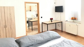 A small, bright room with a bed, a desk, a mirror, a TV, and a window with flowers. - Meeraner Parkresidenz (Meerane)