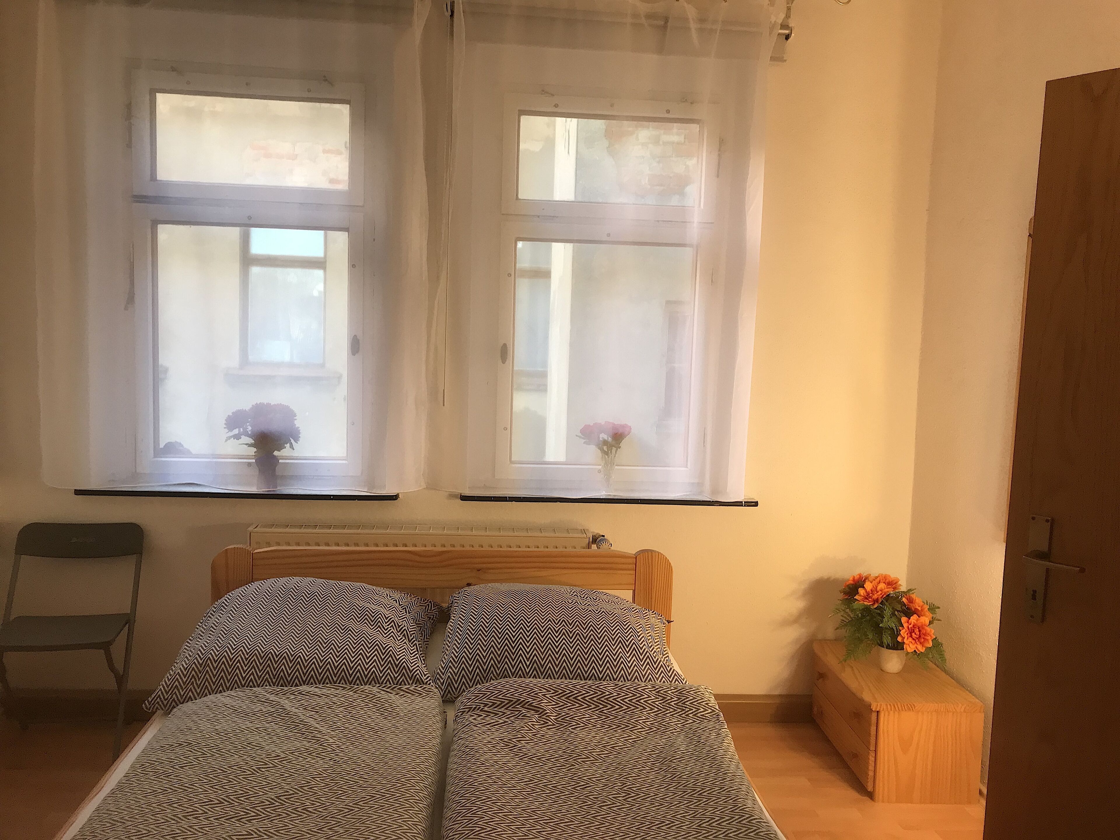 A bedroom with a bed, two windows with curtains, a chair, a bedside table with a flower vase, and a wooden door.