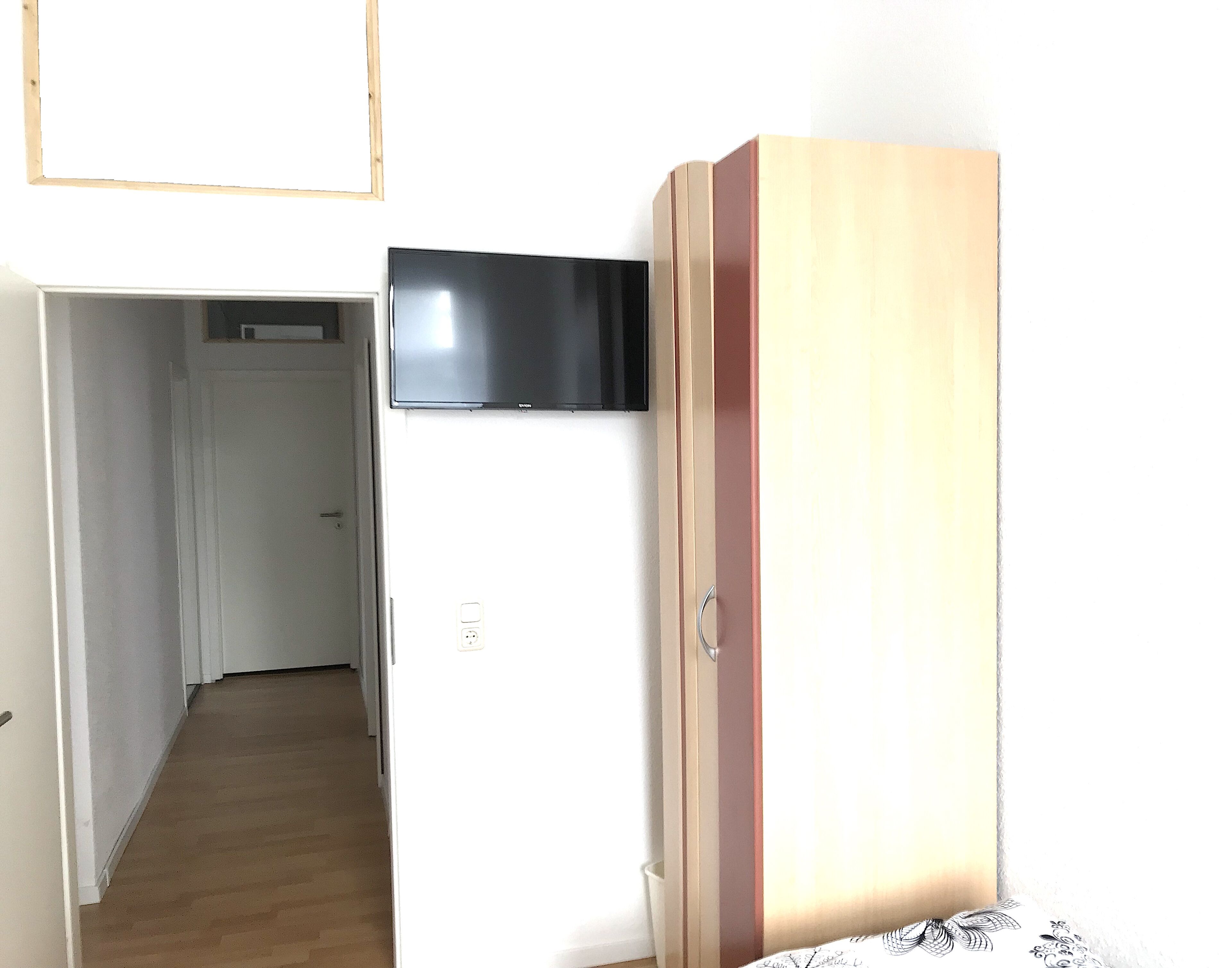 A room with a wooden wardrobe, a television mounted on the wall, and a door leading to another room.