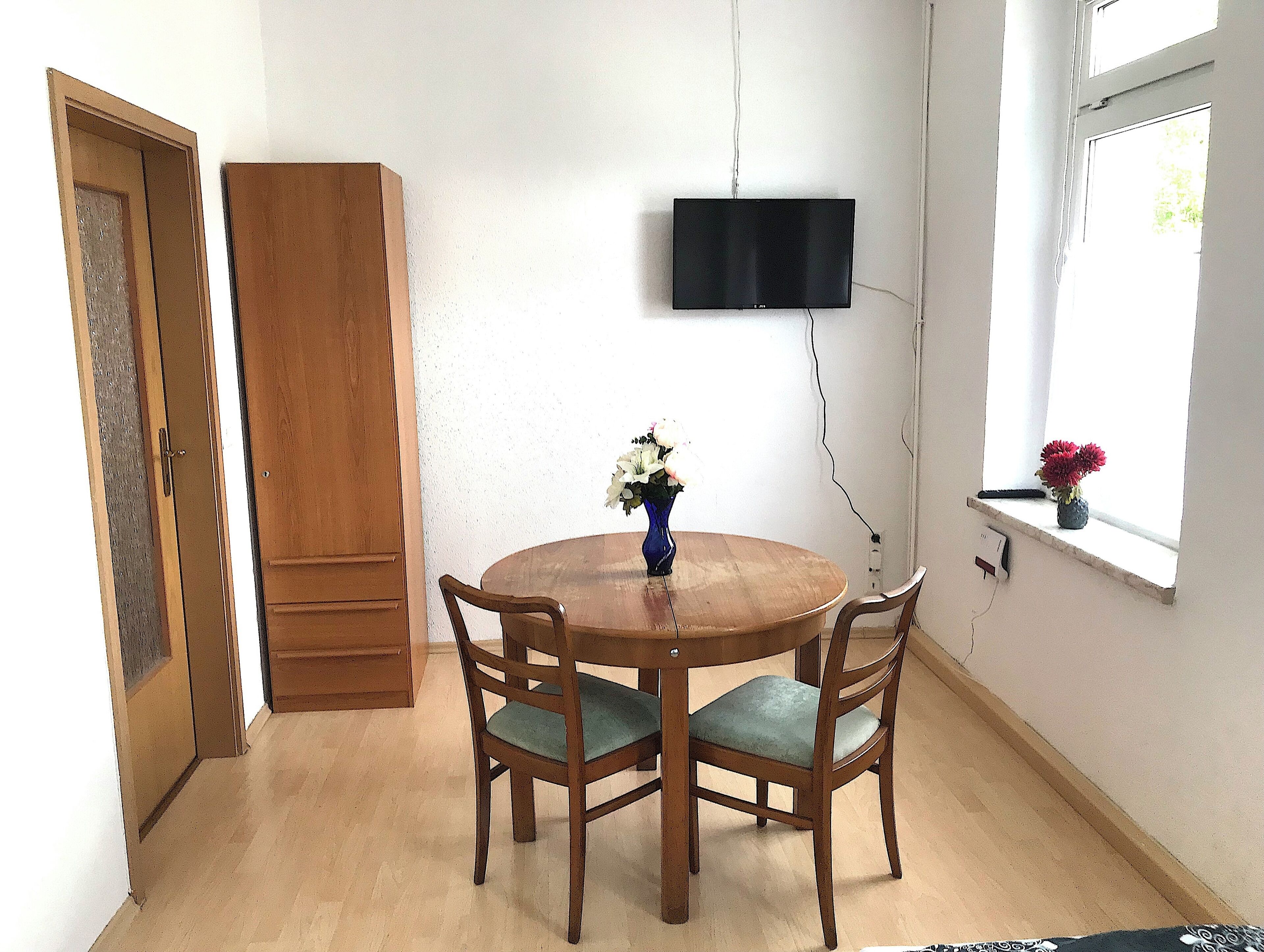 Zona pranzo con tavolo e sedie in legno, un vaso con fiori, un televisore montato a parete e una finestra con una pianta.