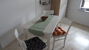 A dining area with a white table, green table runner, and a bowl. There are white chairs with patterned cushions. A small shelf with flowers and a frying pan hangs on the wall.