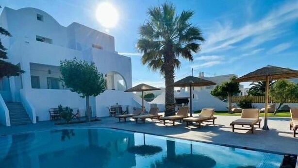 En swimmingpool med liggestole og parasoller, omgivet af en hvid bygning med moderne arkitektur.