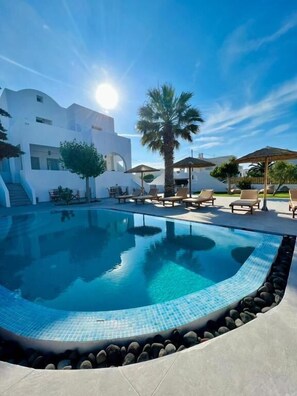 A swimming pool with lounge chairs and umbrellas, surrounded by a white building with a modern design. - Santorini Villatzio Suites (Santorini)