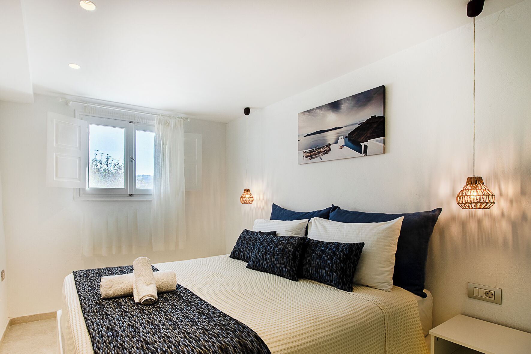Ein Schlafzimmer mit einem Bett, einem Fenster mit Vorhängen, einem Bild an der Wand und zwei Pendelleuchten.