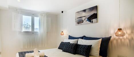 Ein Schlafzimmer mit einem Bett, einem Fenster mit Vorhängen, einem Bild an der Wand und zwei Pendelleuchten.